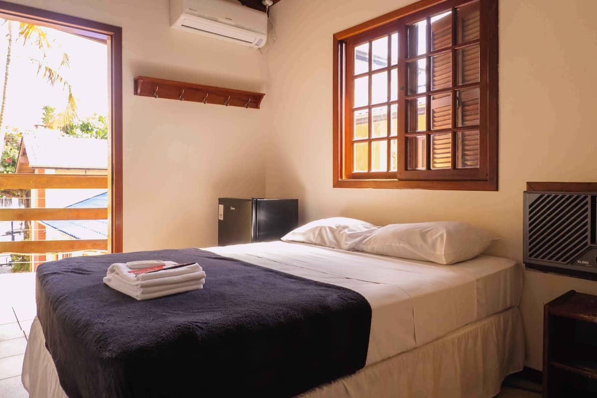 A hotel room with a bed, a window with wooden shutters, a small desk, and a wall-mounted air conditioner.