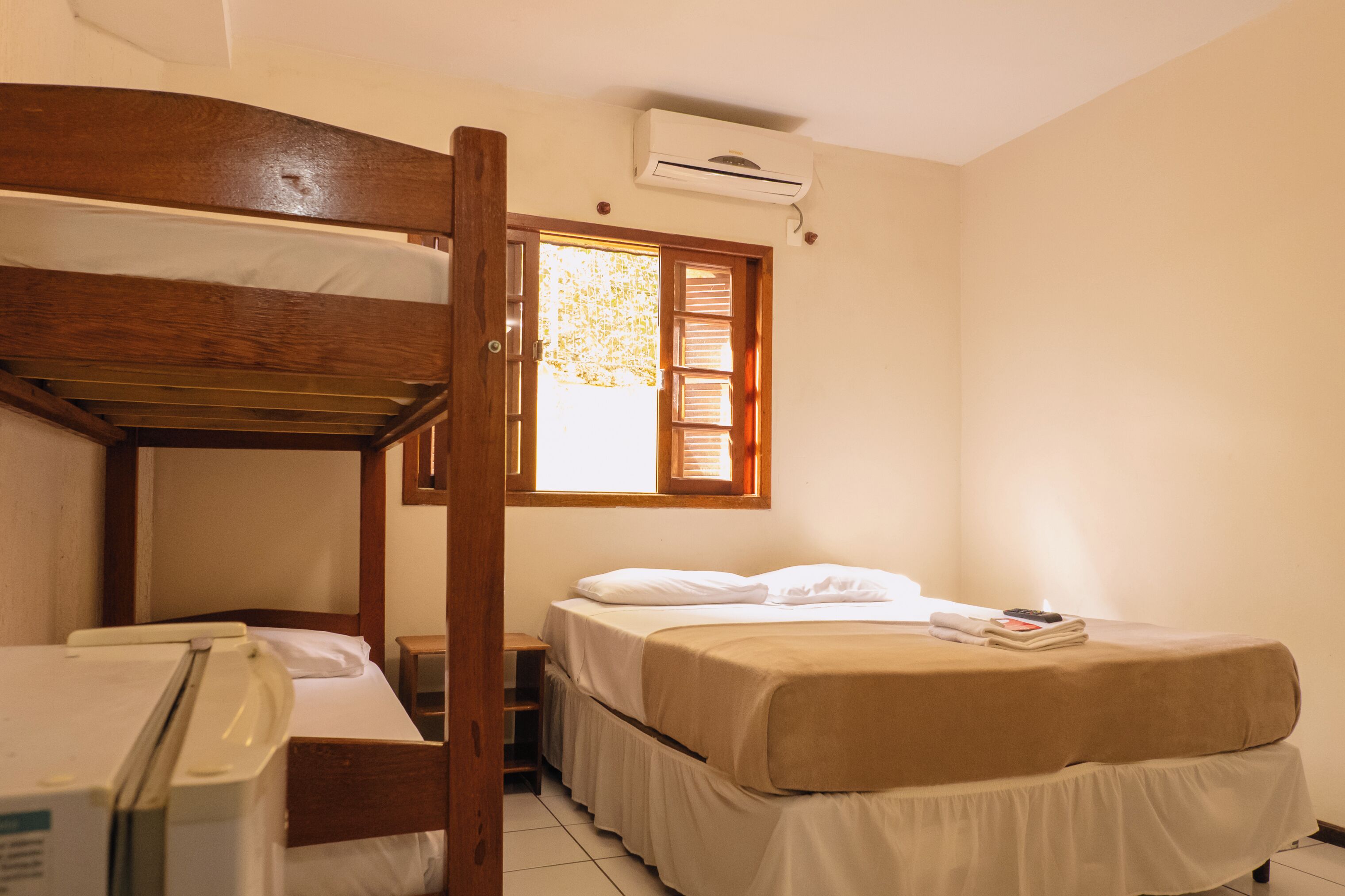 A room with a bunk bed, a single bed, a small desk with a chair, a window with blinds, and an air conditioning unit.