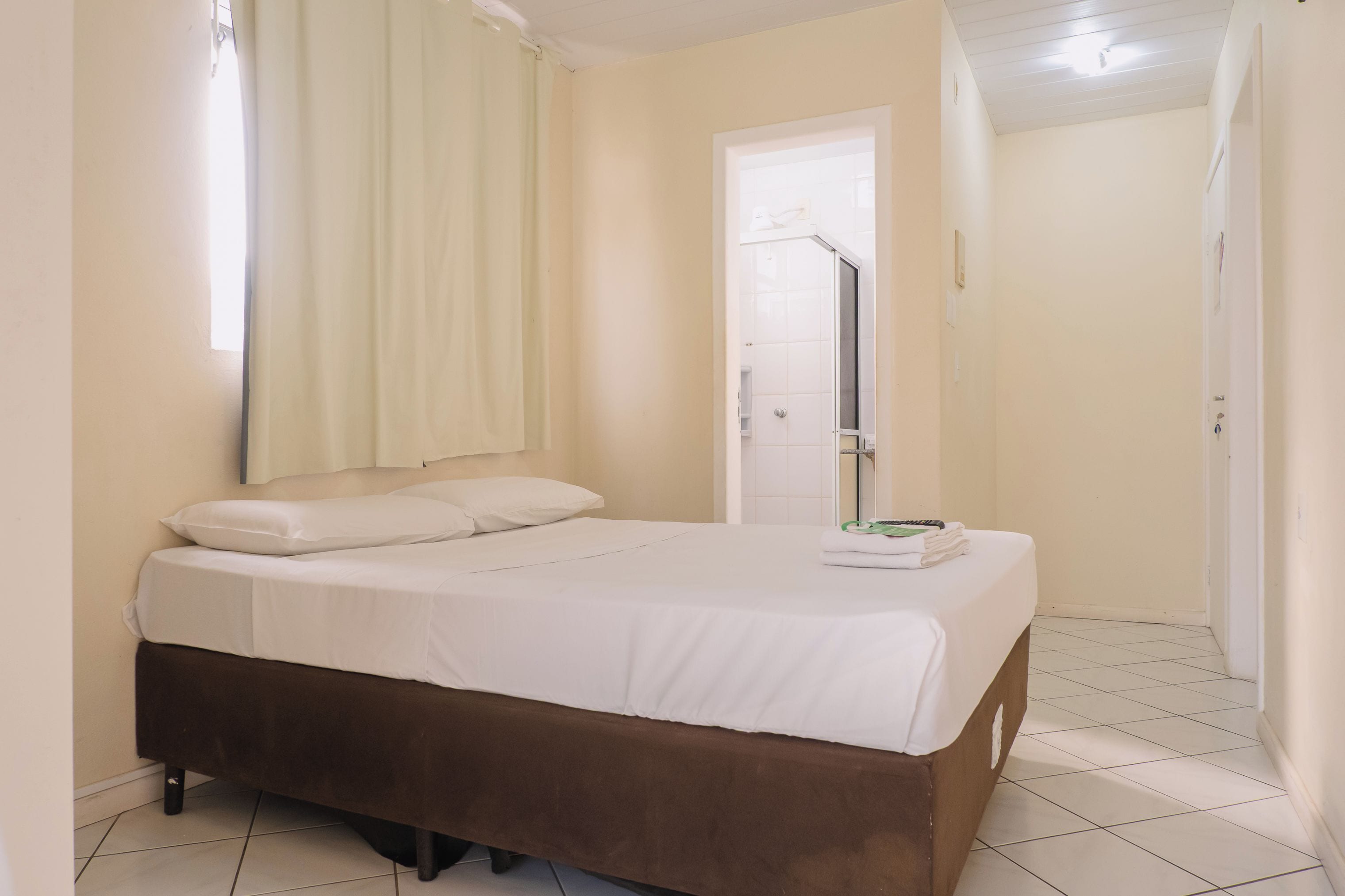 Uma cama de solteiro com cabeceira marrom em quarto com paredes brancas e piso de azulejos.
