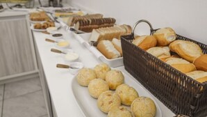 A buffet table with various baked goods including bread, pastries, and small round items, likely cheese balls, arranged on a white table with a wicker basket. - HANNA Amsterdam Hotel & Events (Florianópolis)