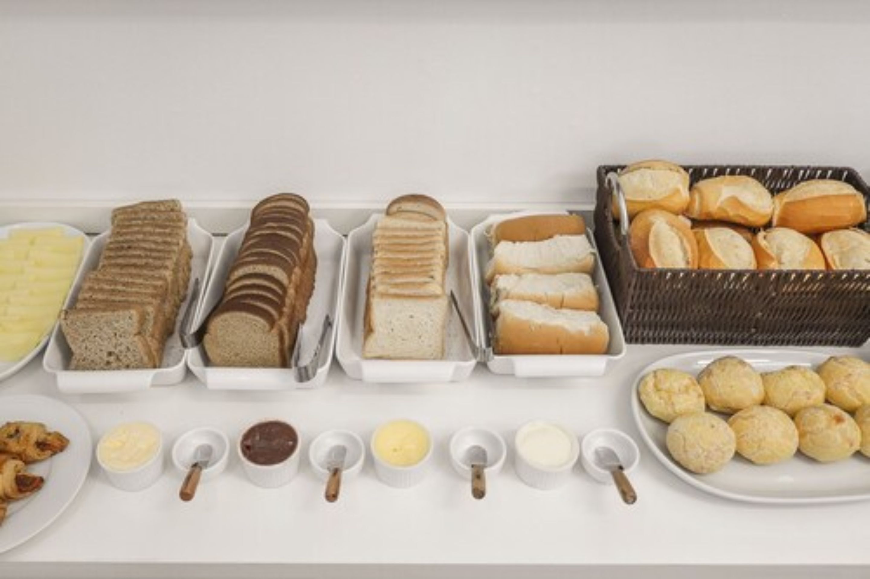 A buffet with various bread and pastries, including sliced bread, rolls, and croissants, along with small bowls of spreads.