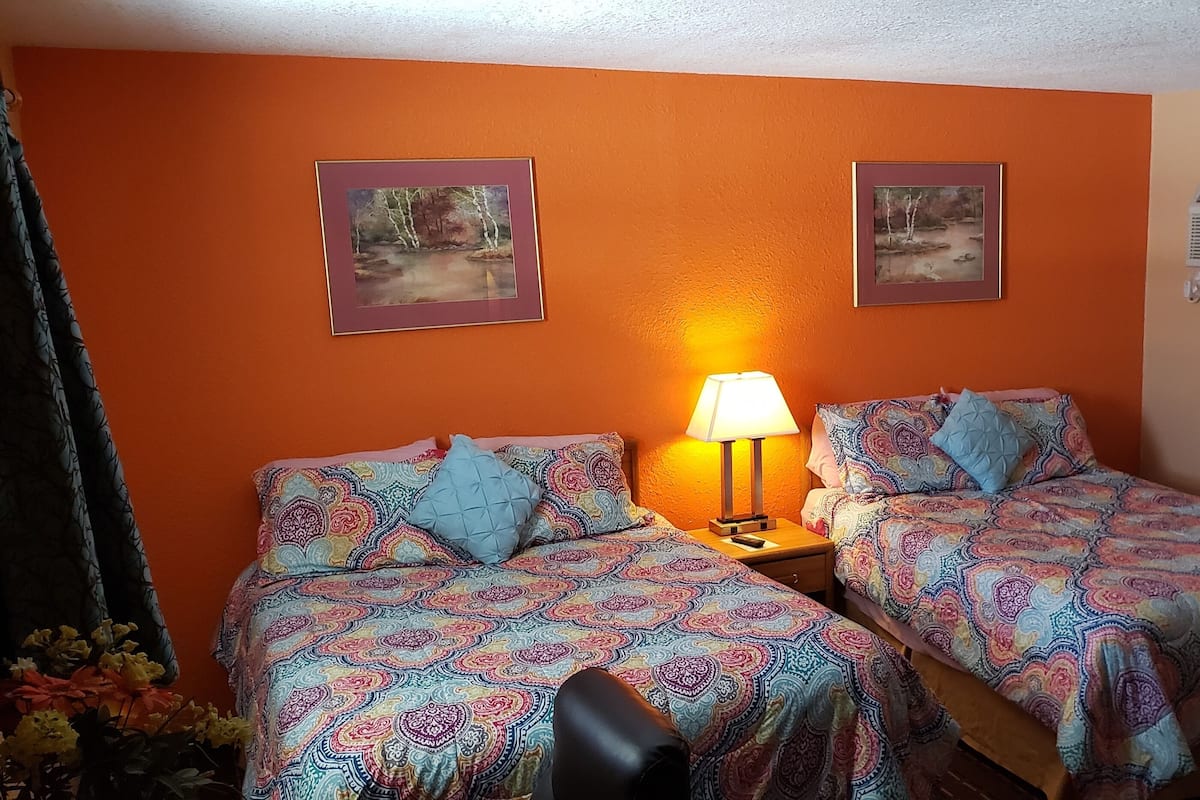 A room with two beds, each with patterned bedding, a bedside table with a lamp, and two framed pictures on the wall.