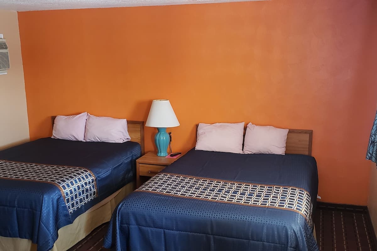 A hotel room with two beds, an air conditioning unit, a nightstand with a lamp, and a window with curtains.