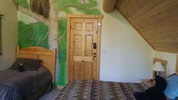 A bedroom with a sloped ceiling, a wooden door, a bed with a patterned bedspread, a nightstand with a lamp, and a wall mural of a bear on a tree branch.