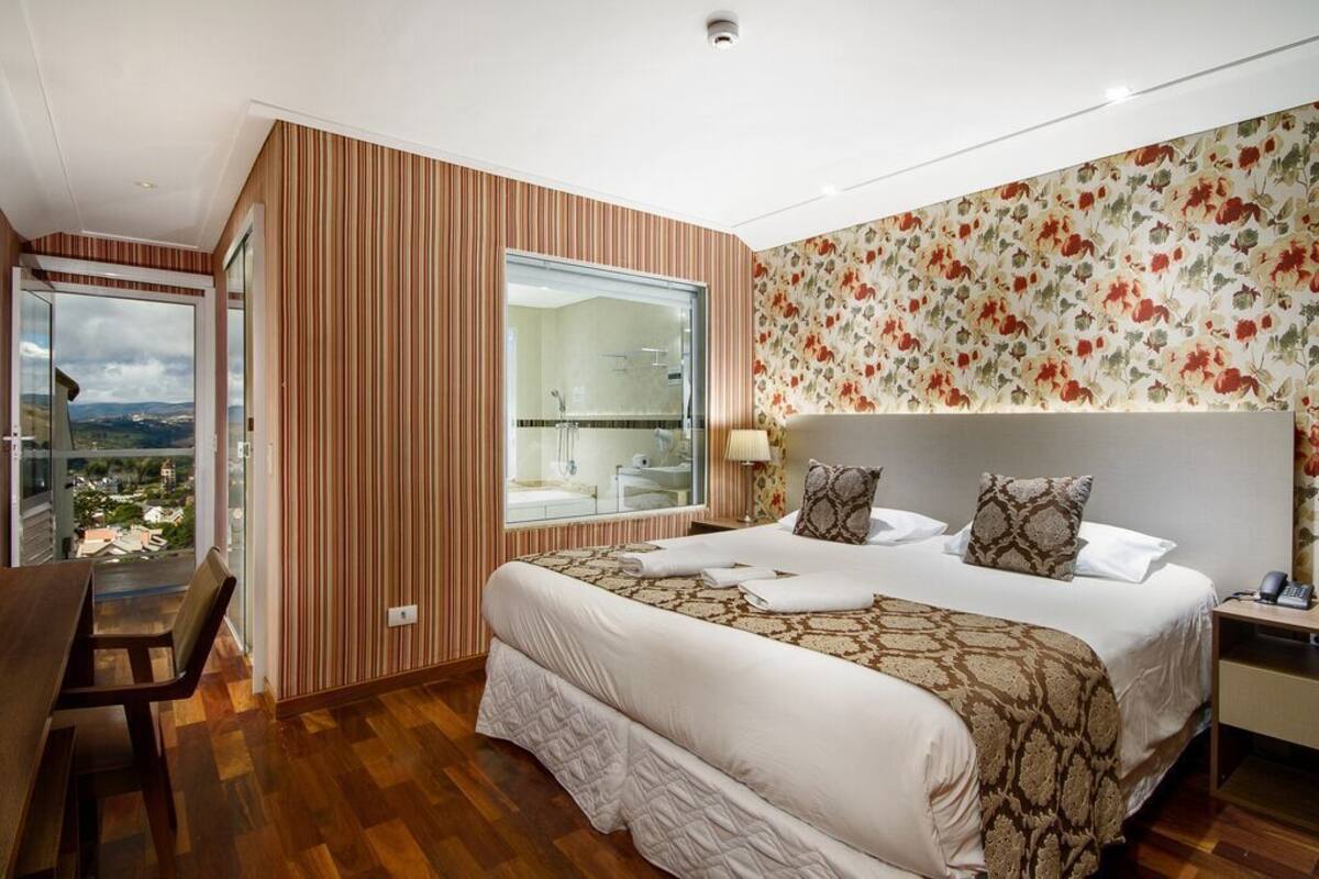 Une chambre avec un grand lit, un bureau, une chaise, une fenêtre donnant sur un beau paysage et une salle de bain visible à travers cette fenêtre.