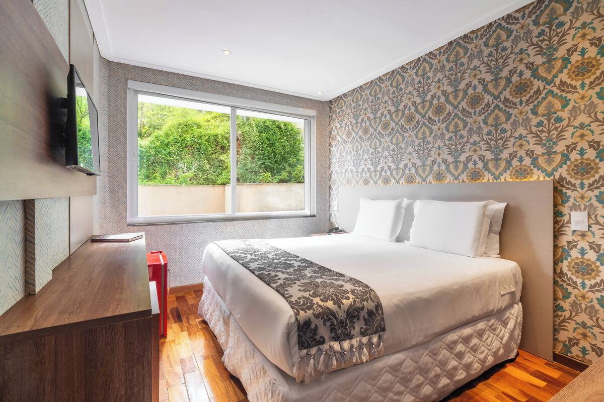 Une chambre moderne avec un grand lit, un téléviseur fixé au mur et une fenêtre donnant sur un paysage verdoyant.