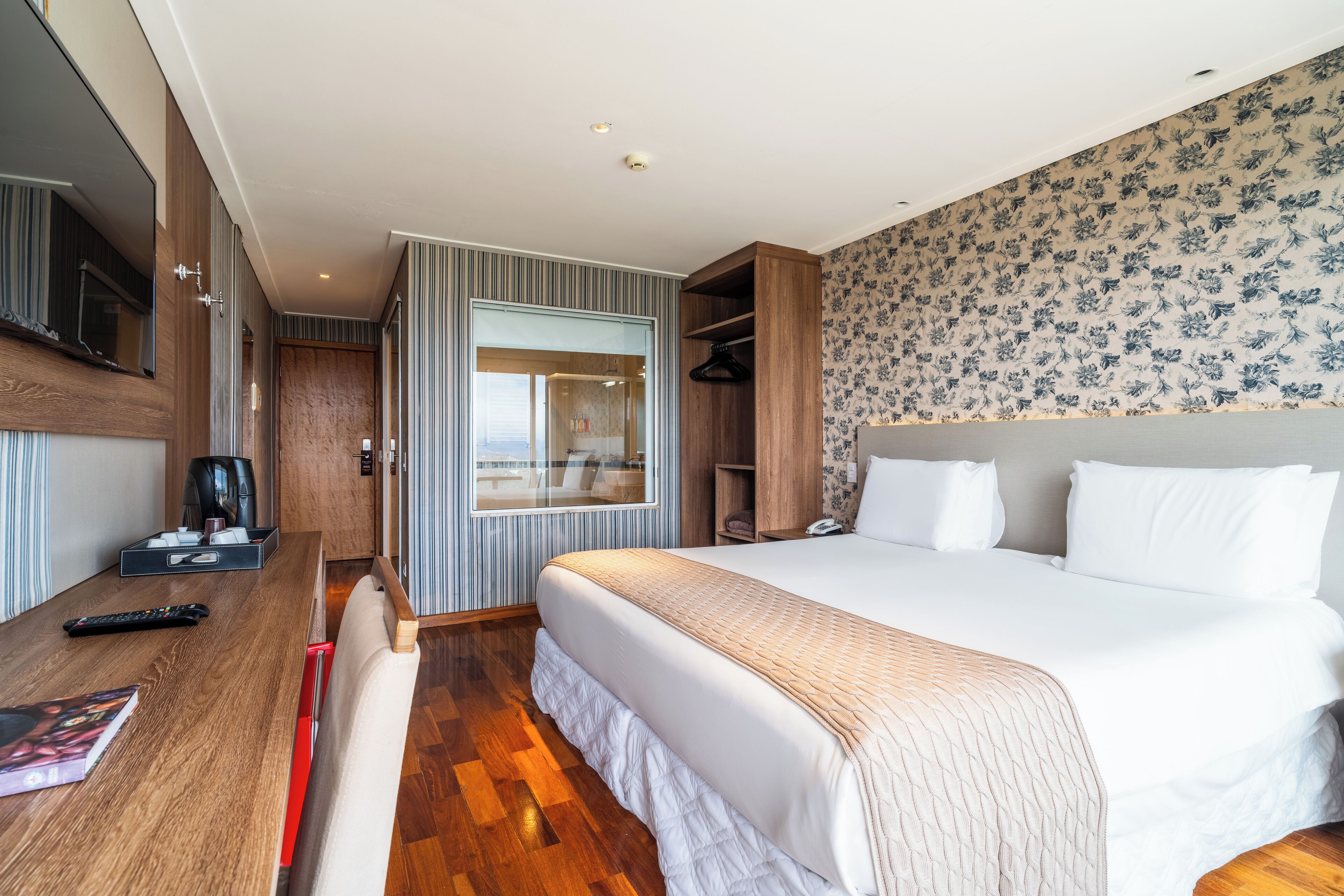 A modern hotel room with a large bed, wooden flooring, a built-in wardrobe, a seating area with a table, and a view through a glass door.