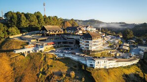 Une vue aérienne d’un hôtel à plusieurs étages, de forme incurvée, entouré d’arbres et avec une chaîne de montagnes en arrière-plan.