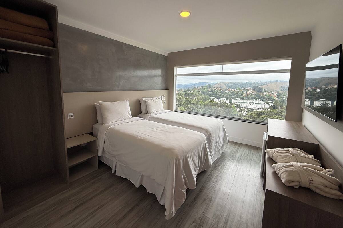 Une chambre d’hôtel moderne avec deux lits, une grande fenêtre donnant sur la ville, une armoire intégrée et un plancher en bois.