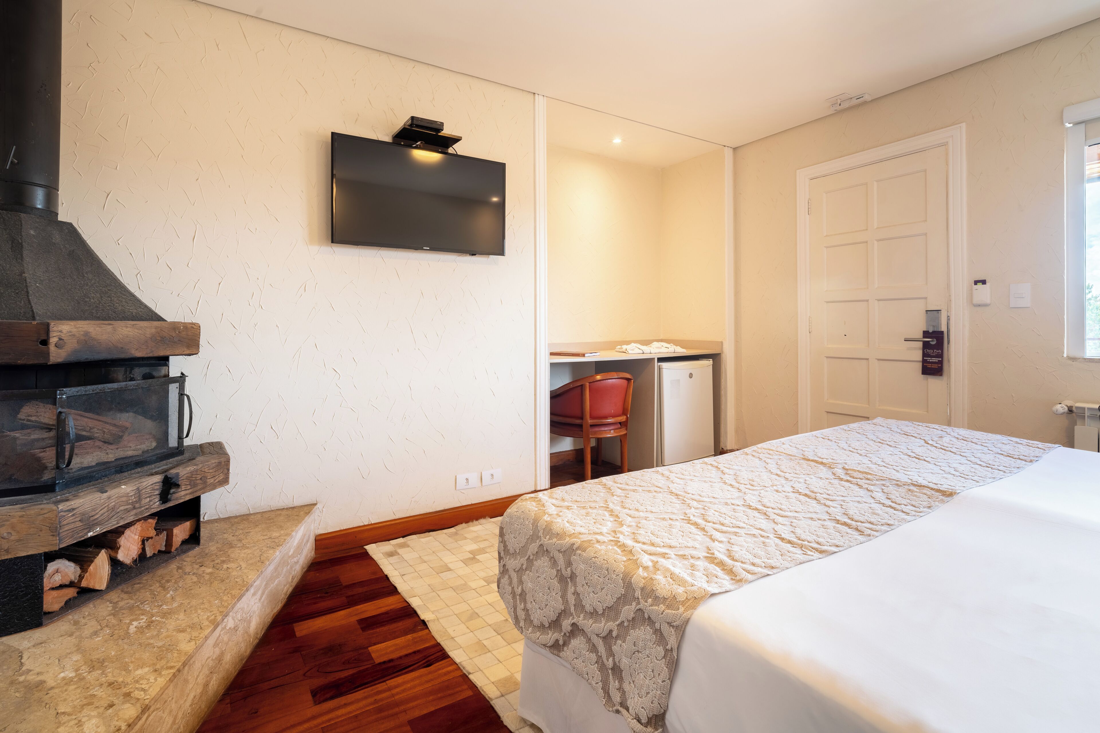 A bedroom with a bed, a TV mounted on the wall, a wooden fireplace, and a door to another room.