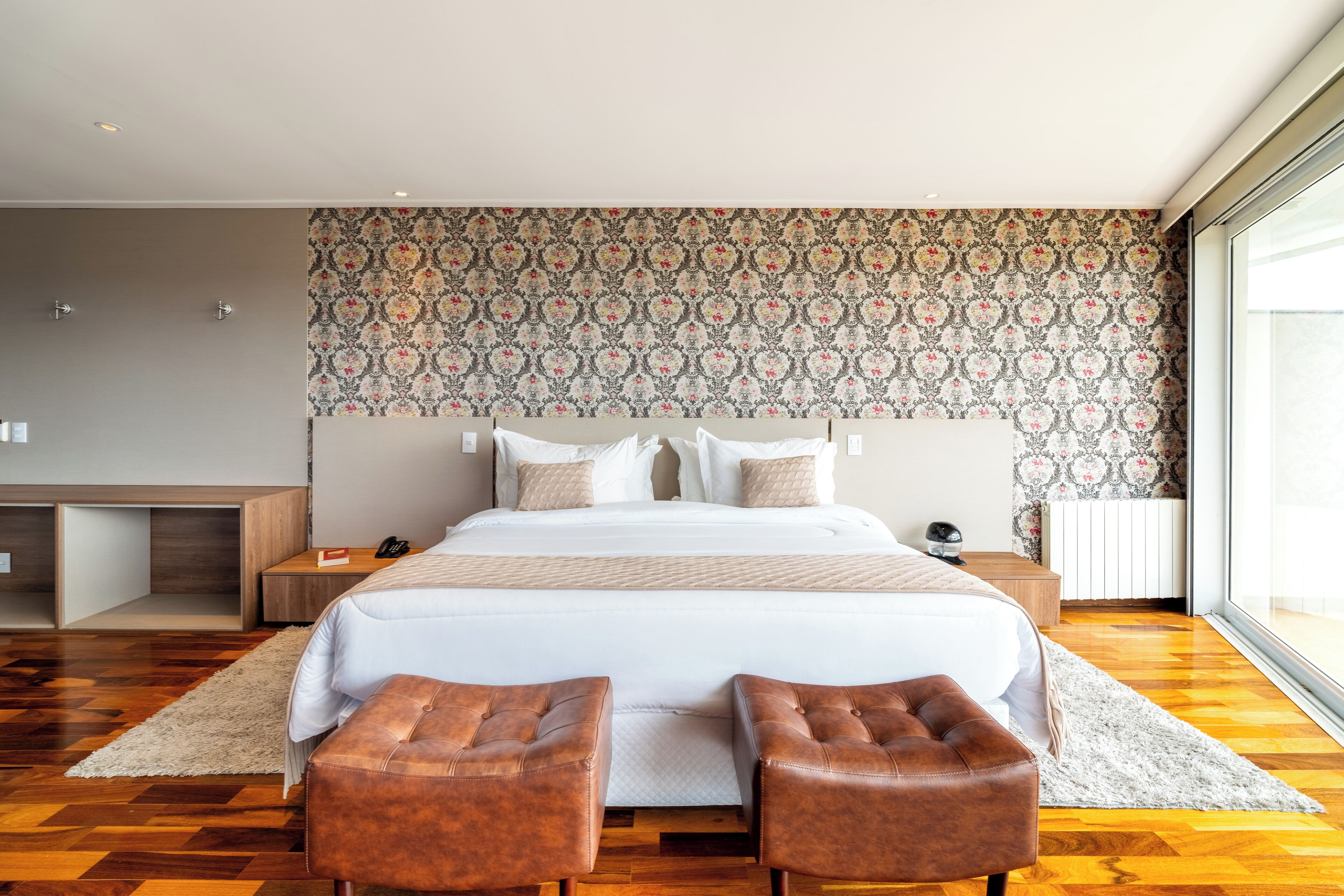 A modern bedroom with a large bed, two brown leather ottomans, a wooden nightstand, and a patterned wallpaper.