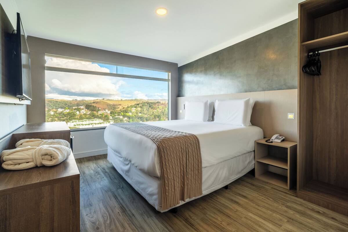 Une chambre moderne avec une grande fenêtre, un lit recouvert d’une couverture beige, une table de chevet avec un téléphone et un banc en bois sur lequel sont posées des serviettes.