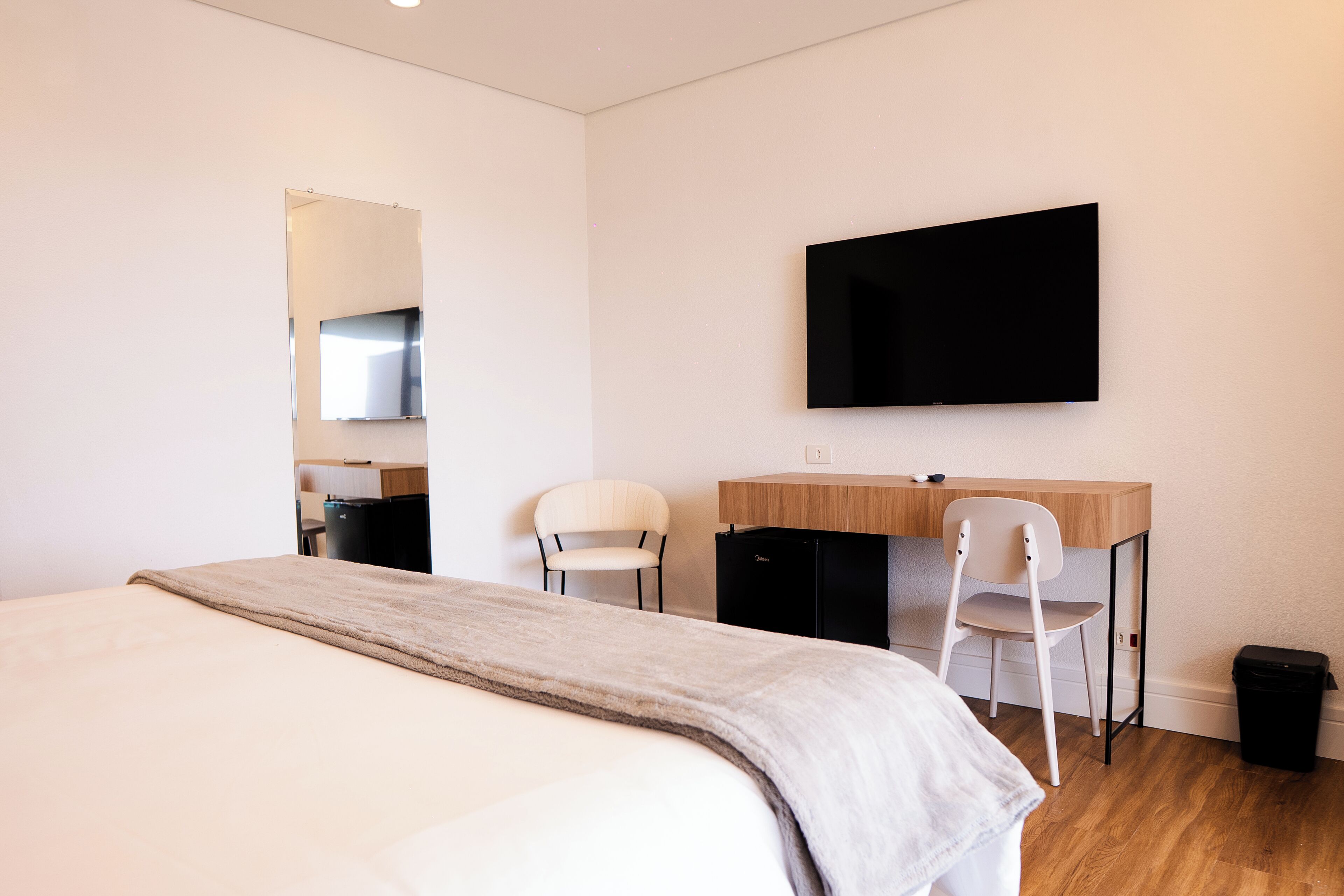 A modern hotel room with a bed, a desk, two chairs, a TV, and a small trash bin.