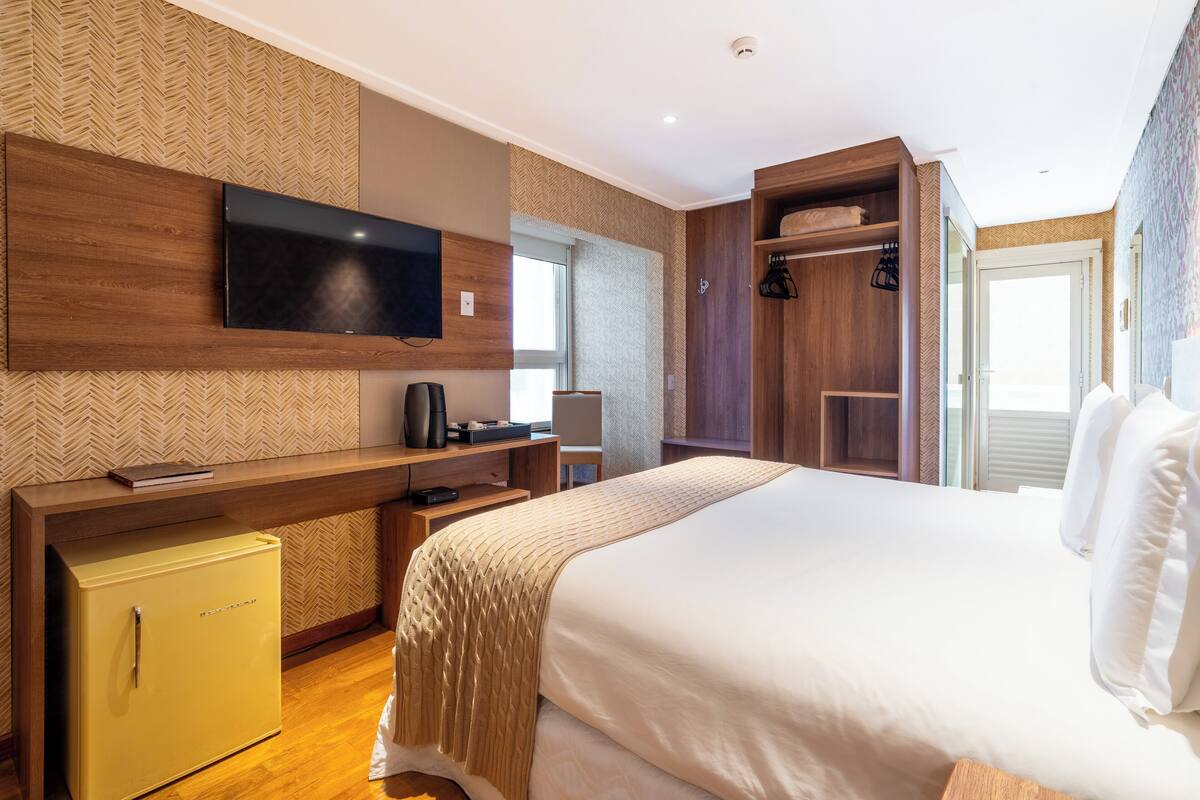 Une chambre d’hôtel moderne équipée d’un lit, d’un téléviseur fixé au mur, d’un petit réfrigérateur jaune et d’un bureau en bois avec une chaise.