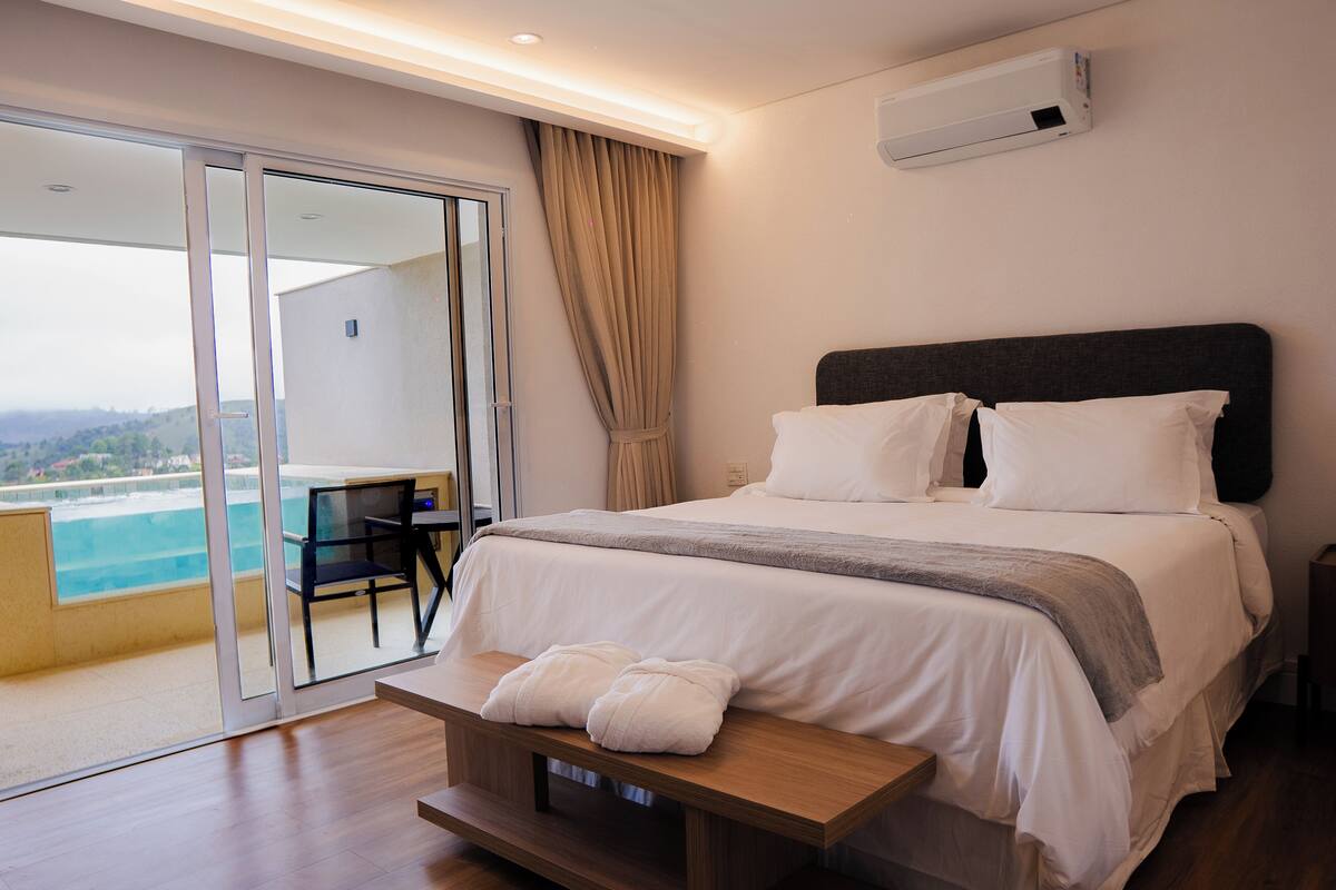 Une chambre d’hôtel avec un lit, une banquette avec des serviettes, un bureau et une vue sur une piscine et des montagnes.