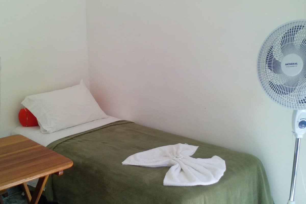 Une petite chambre avec un lit simple, une table en bois, un oreiller rouge, une serviette blanche sur le lit et un ventilateur sur pied.