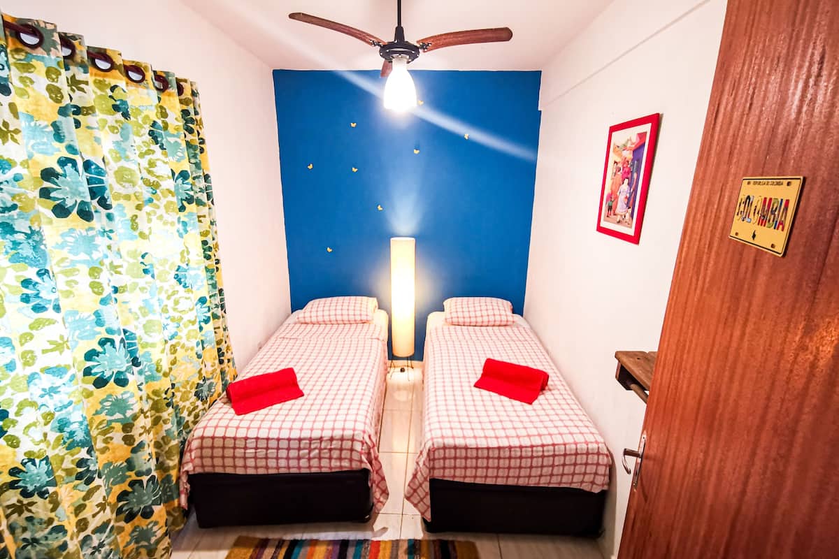Une chambre avec deux lits, un ventilateur de plafond, une table de chevet, une lampe, un tapis rayé aux couleurs vives et un mur orné d’un tableau et d’une plaque.
