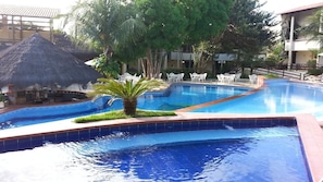 A swimming pool with a thatched-roof structure, surrounded by palm trees and white chairs. - Solar De Pipa Duplex Flat (Tibau do Sul)