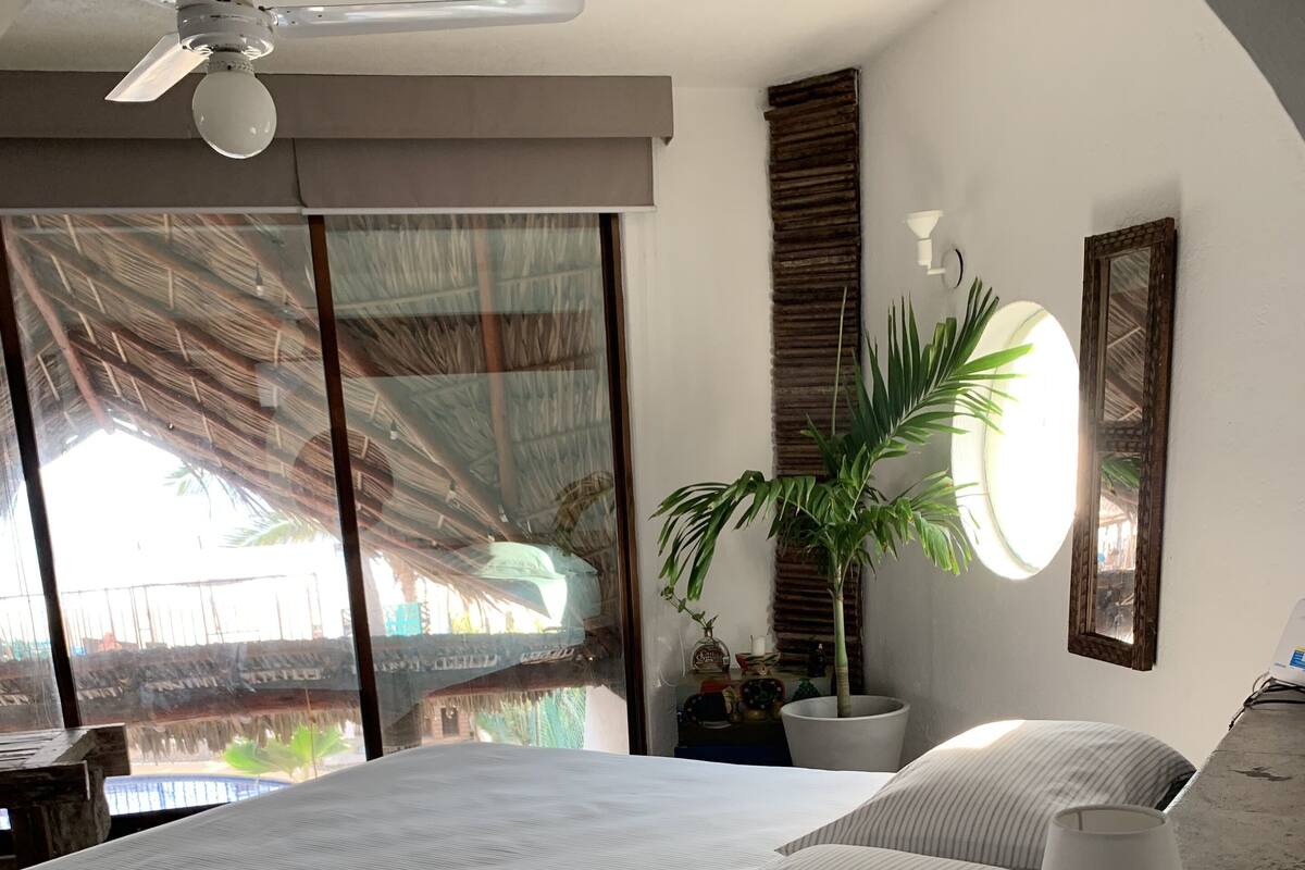 Un dormitorio con una cama grande, un ventilador de techo, una planta en maceta y vista a una construcción con techo de paja.