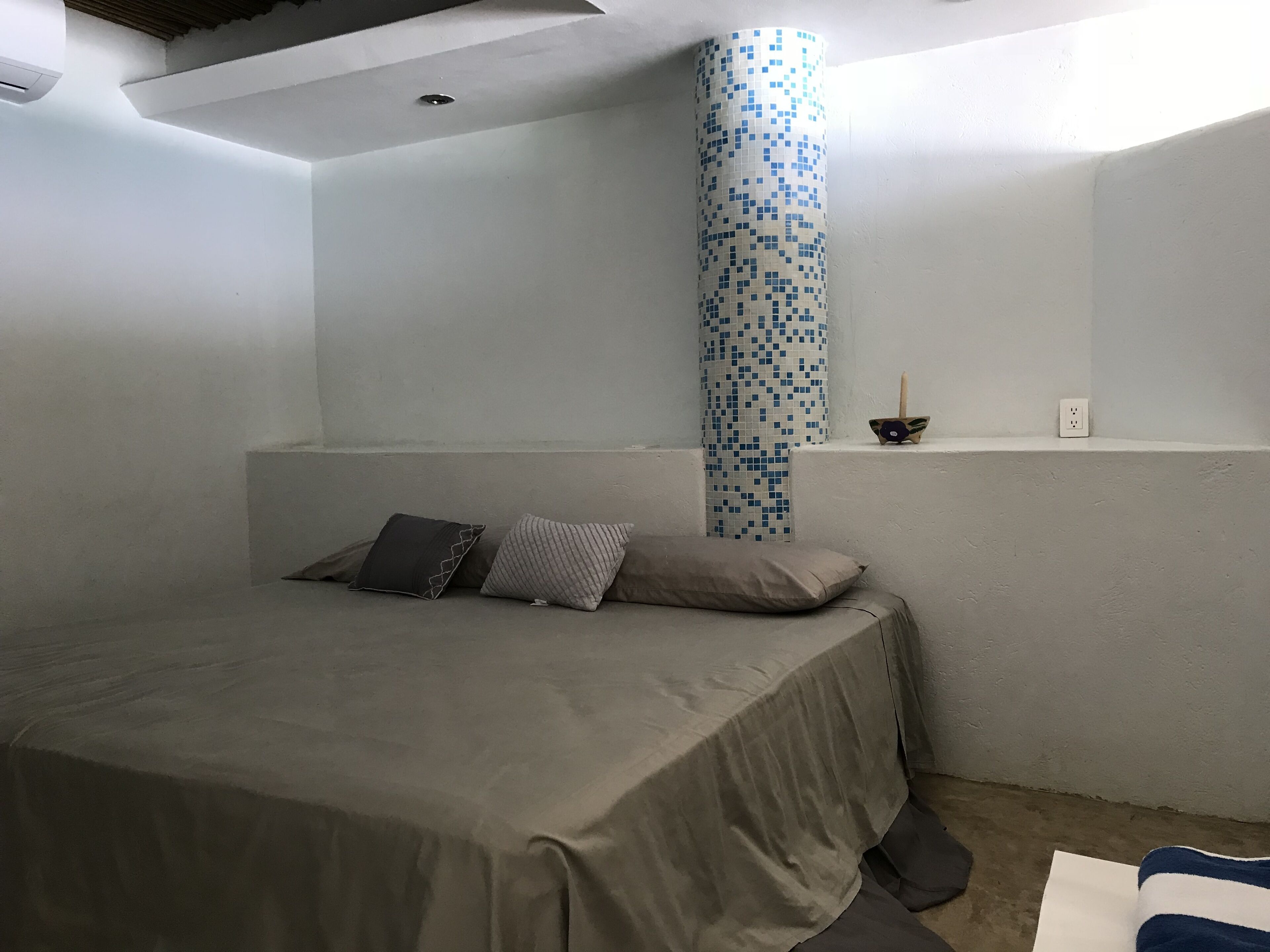 Une chambre à coucher au style minimaliste, avec un lit, des oreillers, une petite étagère et une bouche d’aération fixée au mur.