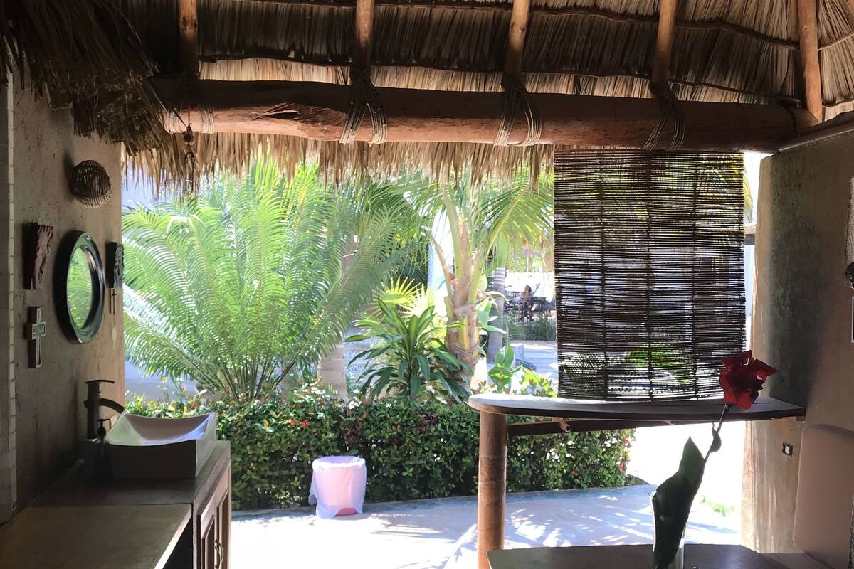 Una habitación con techo de paja, un lavabo, un espejo y ventana con vistas a un jardín con palmeras.