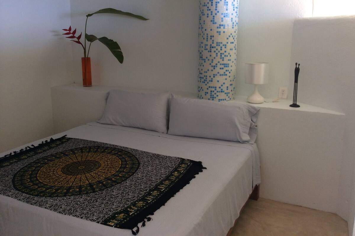 Un dormitorio con cama, un tapiz decorativo, una planta en maceta, una lámpara de pared y una pared con diseño azul y blanco.