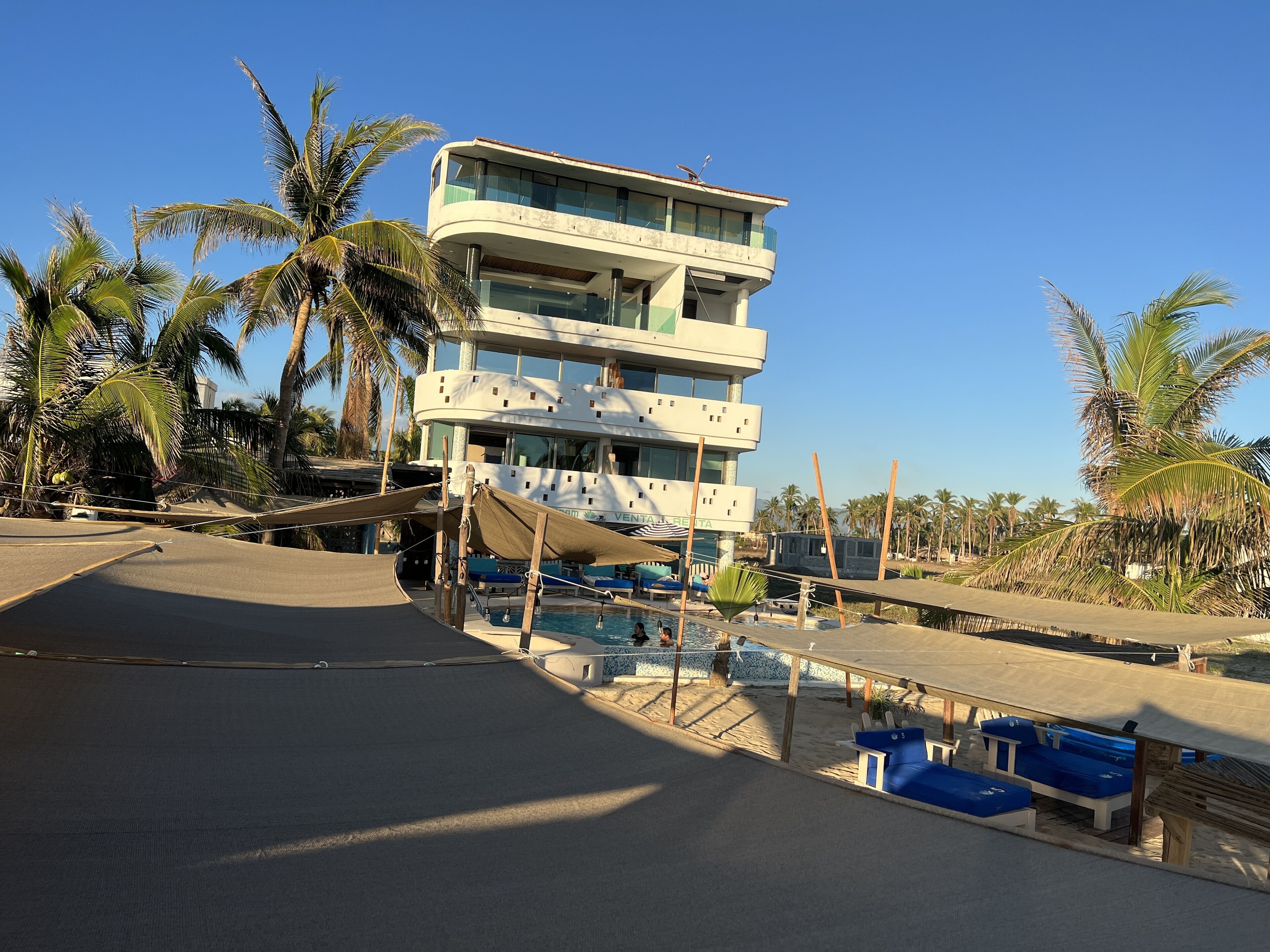 A multi-story hotel with a pool area, palm trees, and a clear blue sky.