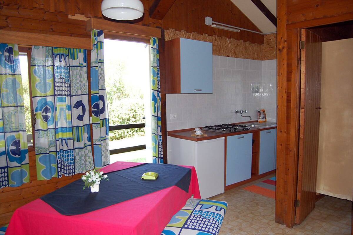 A compact kitchen with a gas stove, sink, and cabinets, a dining area with a table and chairs, and a view through a window with patterned curtains.