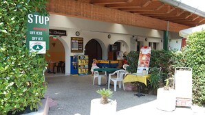 An outdoor area with a 'STOP' sign, a 'UFFICIO OFFICE' sign, and a 'CAMPEGIO' sign. There are tables and chairs, and a small kiosk with various items.