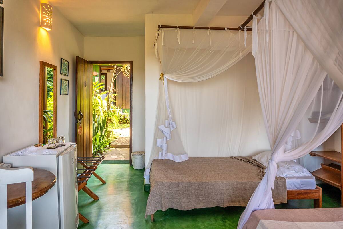 A room with a bed, a small table, and a bench. There is a door leading to a garden.