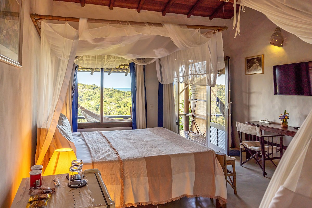 A bedroom with a bed, a desk, and a chair. There is a window with a view of greenery.