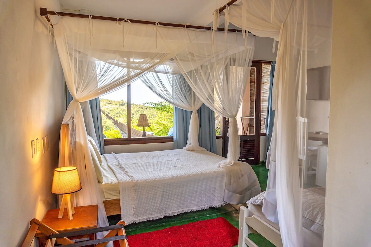 A bedroom with a bed under a canopy, a bedside table with a lamp, and a view of greenery outside.