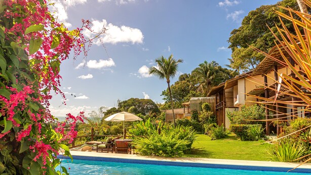 A swimming pool with a tiled edge, surrounded by lush greenery and a wooden structure. - Pousada Mar à Vista (Porto Seguro)