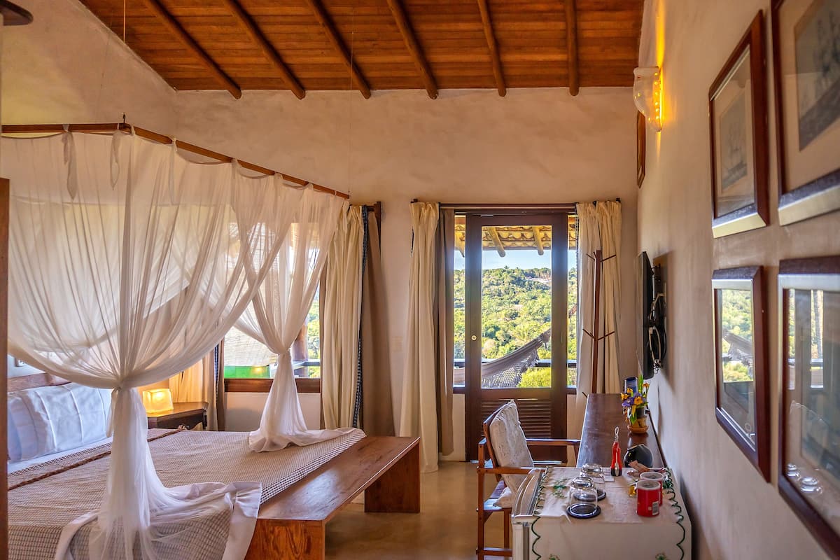 A room with a bed, a wooden table with a chair, and a view of greenery outside.