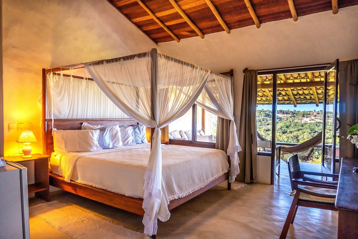 A four-poster bed with a canopy, a bedside table with a lamp, and a view of a lush landscape through a large window.