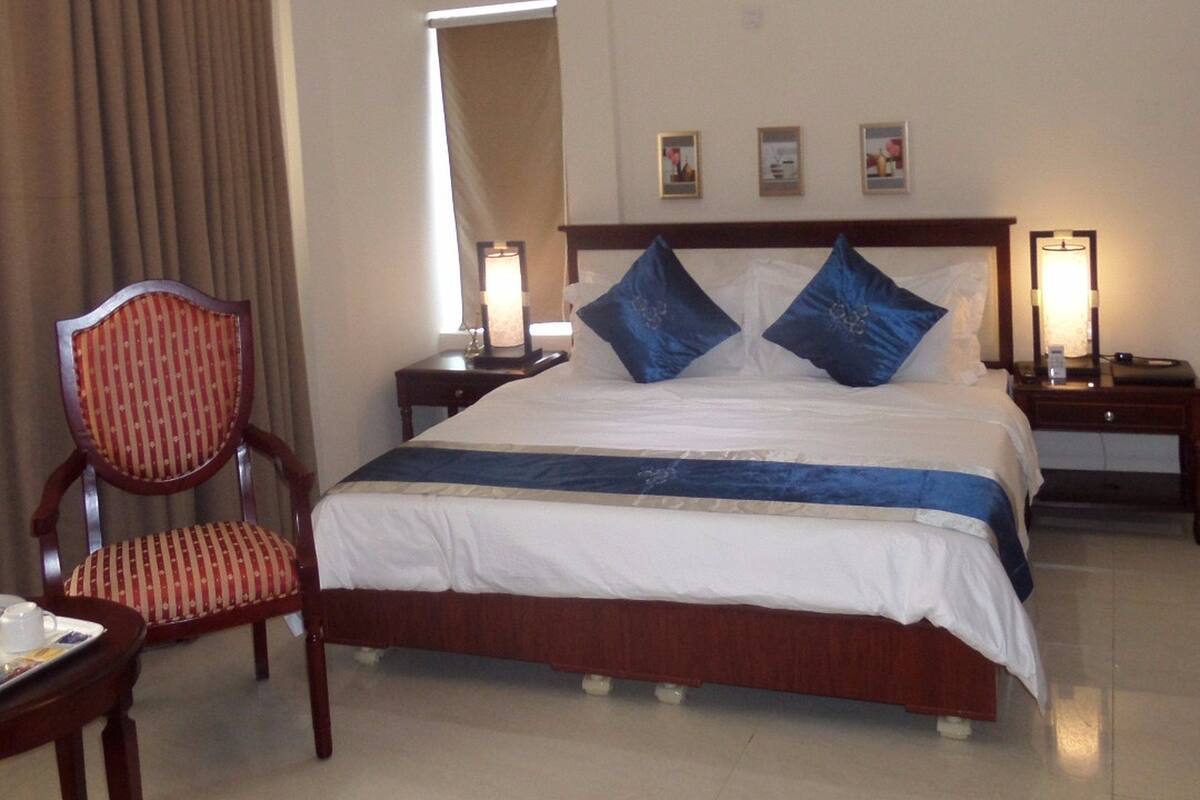 A hotel room with a bed, a chair, a bedside table, a nightstand, and a wall-mounted air conditioning unit.