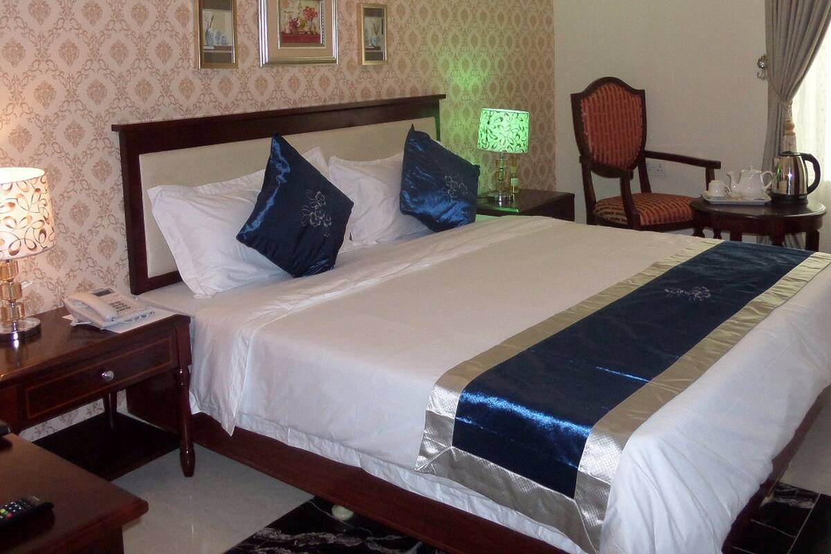 A hotel room with a large bed, a nightstand with a lamp, a chair, and a desk with a telephone.