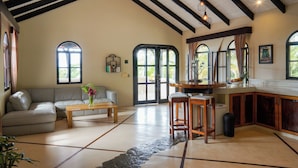 A spacious living area with a high ceiling, a sofa, a coffee table, and a kitchen area with wooden cabinets and a window. - Casa Marbella (Cóbano)