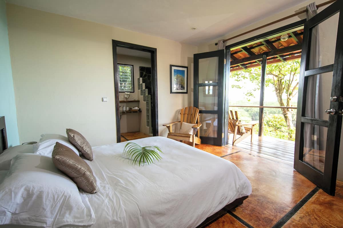 A bedroom with a large bed, a wooden chair, and a balcony with a view of greenery.