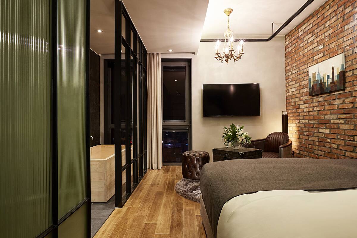 A modern hotel room with a large bed, a flat-screen TV mounted on the wall, a brick accent wall, and a view of the cityscape through the window.