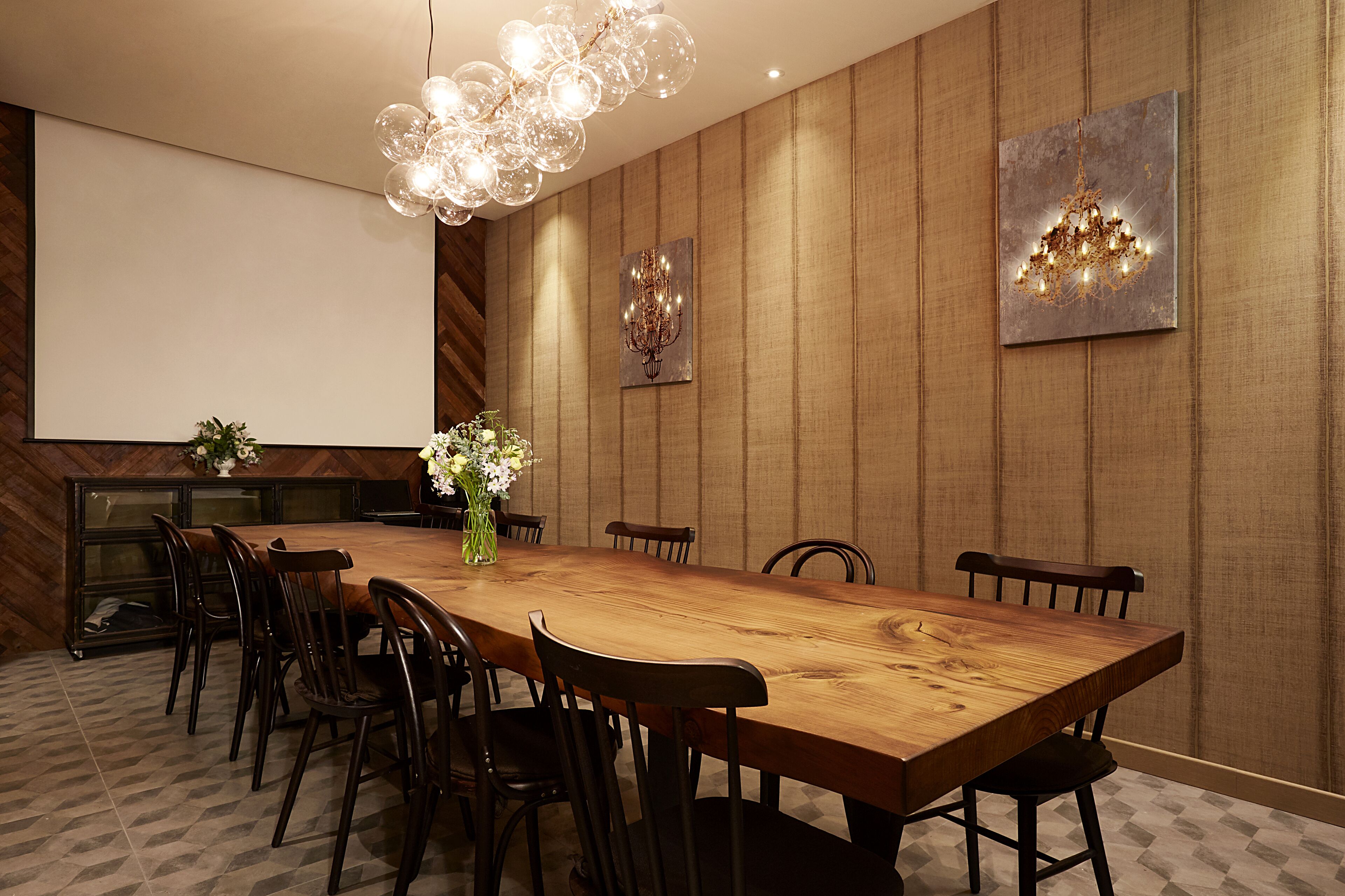 A conference room with a long wooden table, multiple chairs, a projector screen, and two chandeliers.