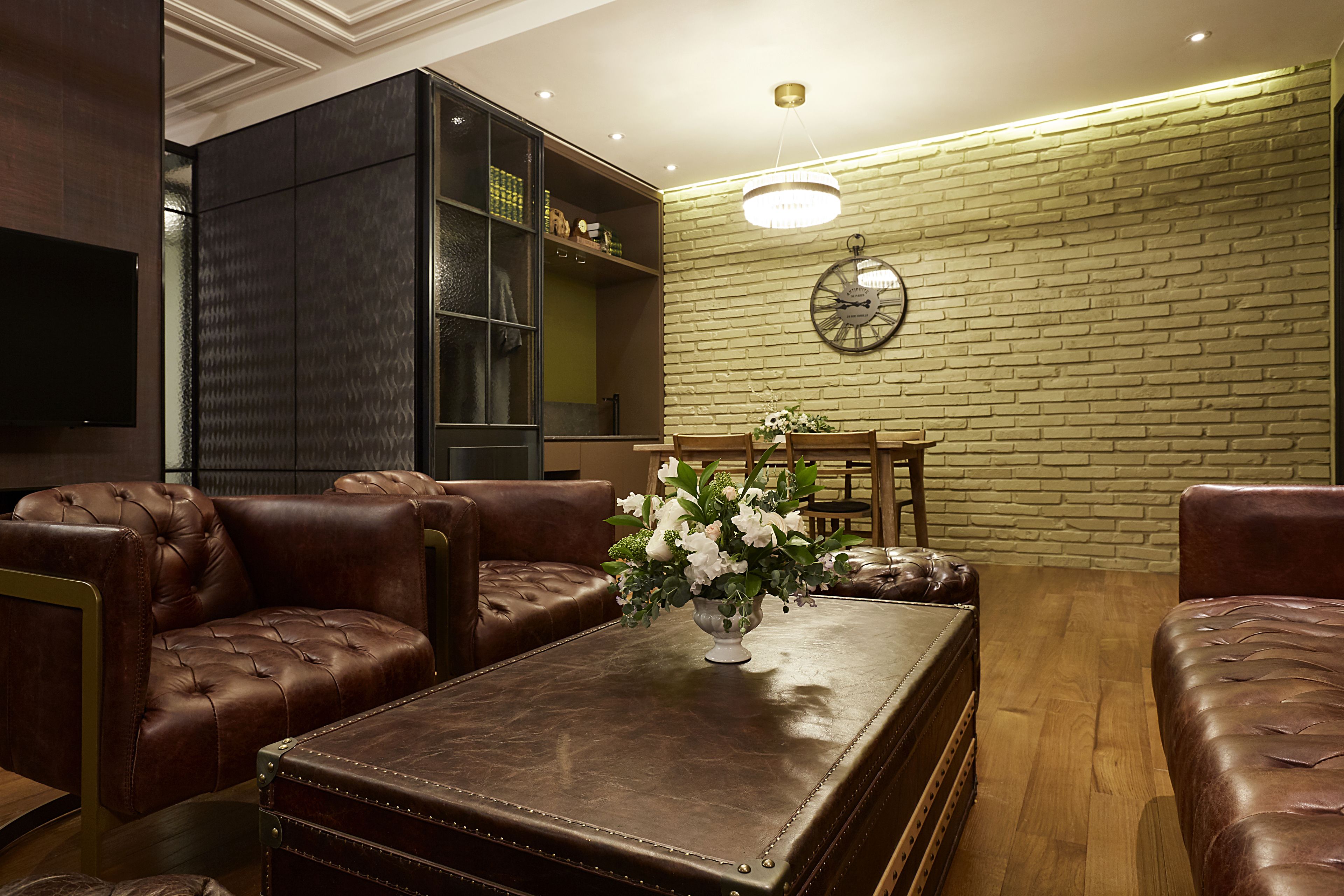 Un salon moderne avec des canapés en cuir, une table basse ornée d’un vase de fleurs, un mur en briques et une horloge murale ronde.
