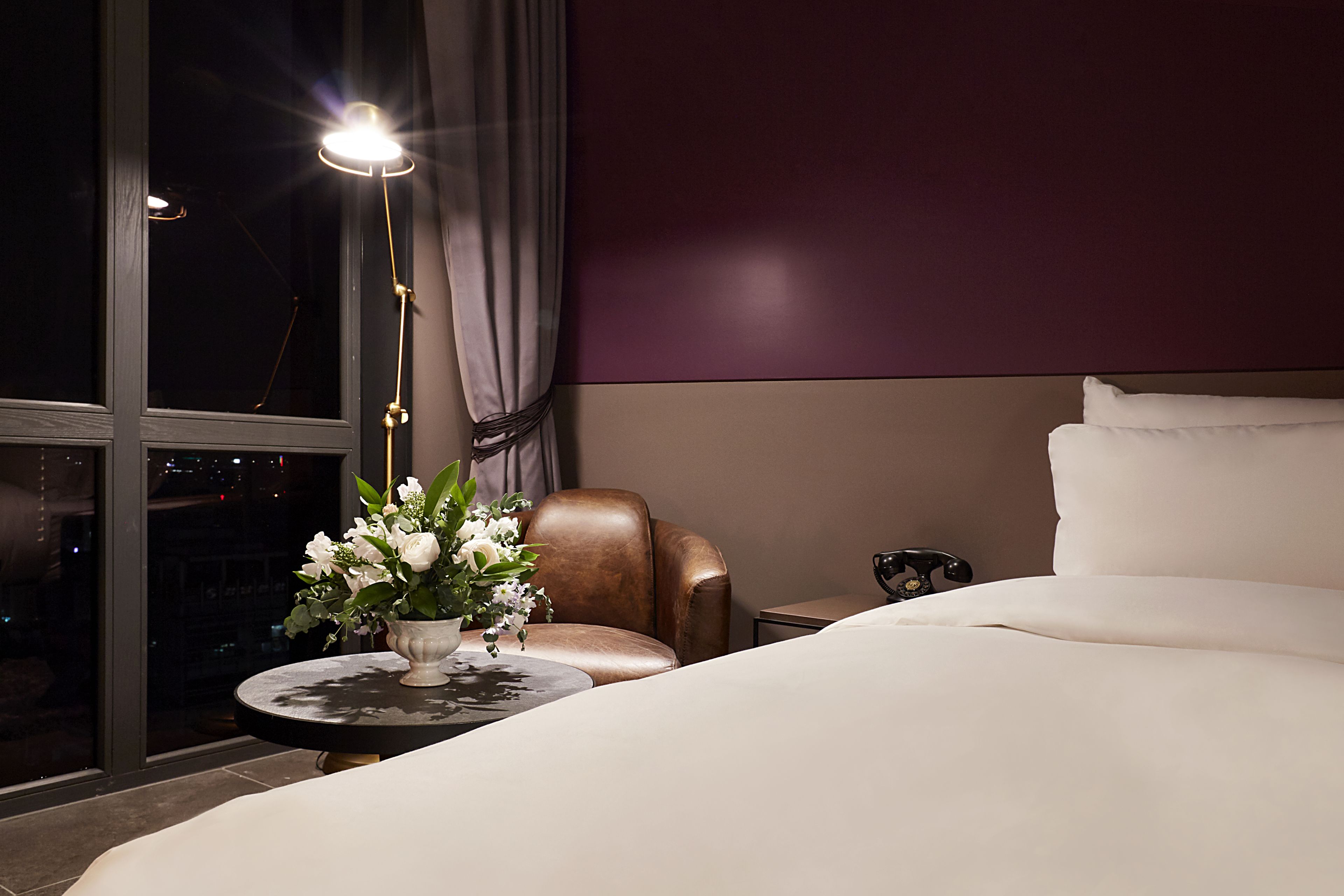 A hotel room with a large bed, a round table with a vase of flowers, a leather armchair, and a nightstand with a lamp.
