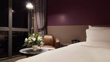 A hotel room with a large bed, a round table with a vase of flowers, a leather armchair, and a nightstand with a lamp.