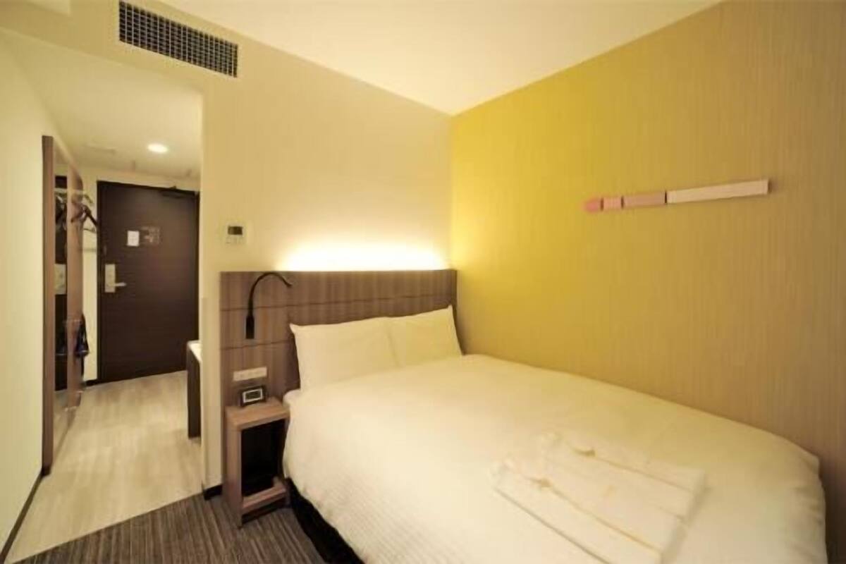 A hotel room with a bed, bedside table, and a wall-mounted light.