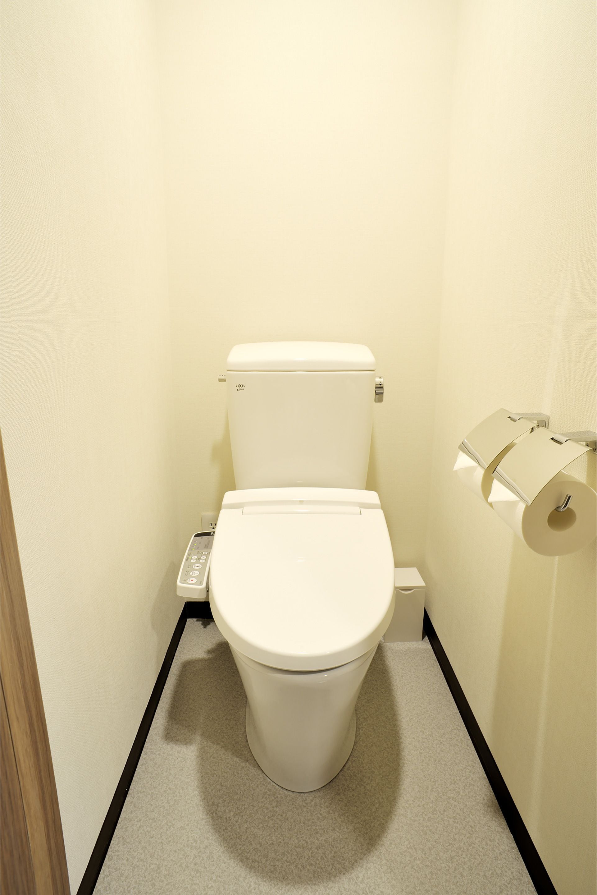 A small, compact bathroom with a white toilet, a toilet paper holder, and a control panel.