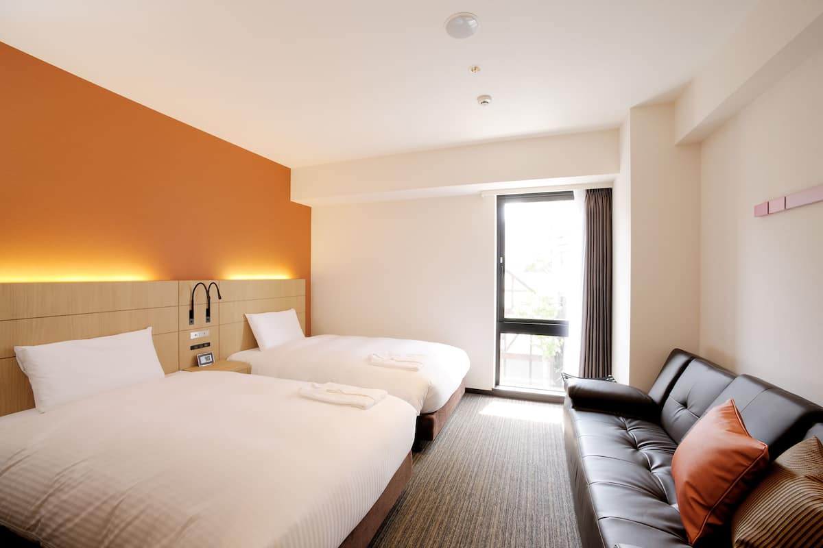 A hotel room with two beds, a sofa, a window, and a door.