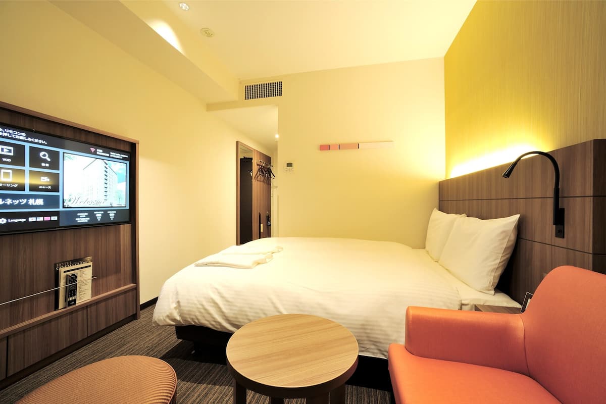 A hotel room with a bed, a TV mounted on the wall, a small round table, and an orange chair.