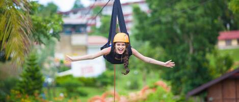 A person suspended in a hammock, with greenery and buildings in the background.