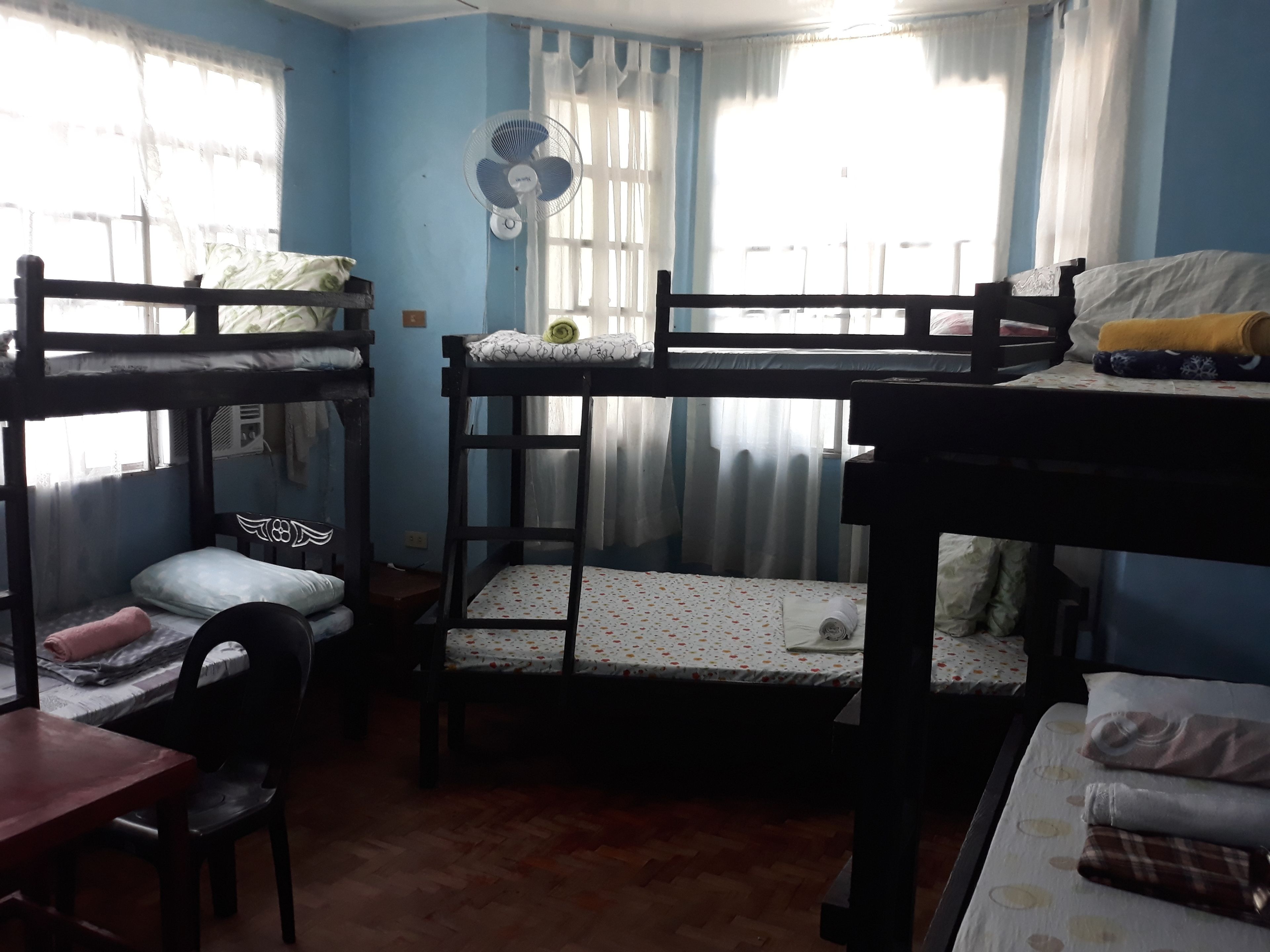 A dormitory room with bunk beds, a desk, a chair, and a fan.